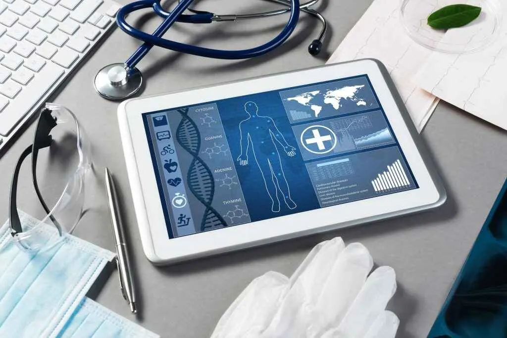 A digital tablet displaying medical data and a human body diagram sits on a desk surrounded by medical equipment including a stethoscope, keyboard, pen, goggles, gloves, and a surgical mask.