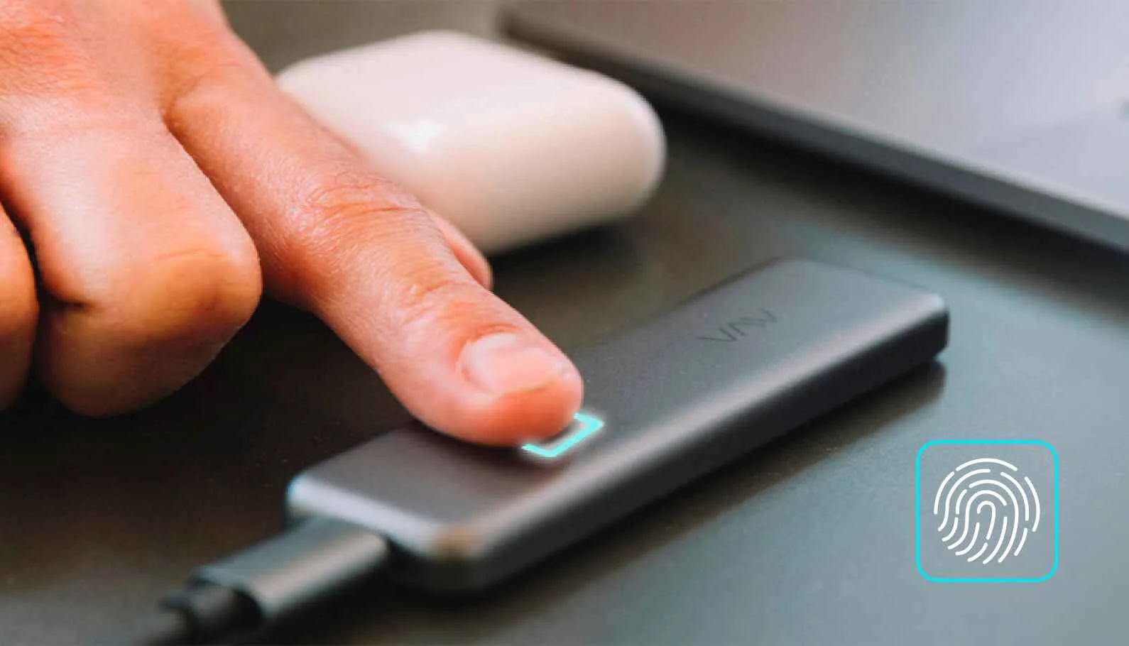 A close-up of a person’s finger touching a fingerprint sensor on a sleek, black electronic device, with a white wireless earbud case and laptop nearby on the desk, illustrating biometric authentication technology.