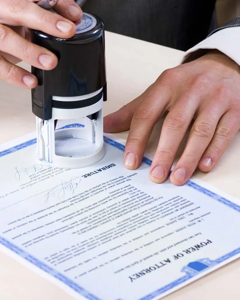 A person is stamping an official document labeled "Power of Attorney" with a notary stamp, indicating the document is being notarized. The person's hands and the stamp device are clearly visible on a light-colored desk.