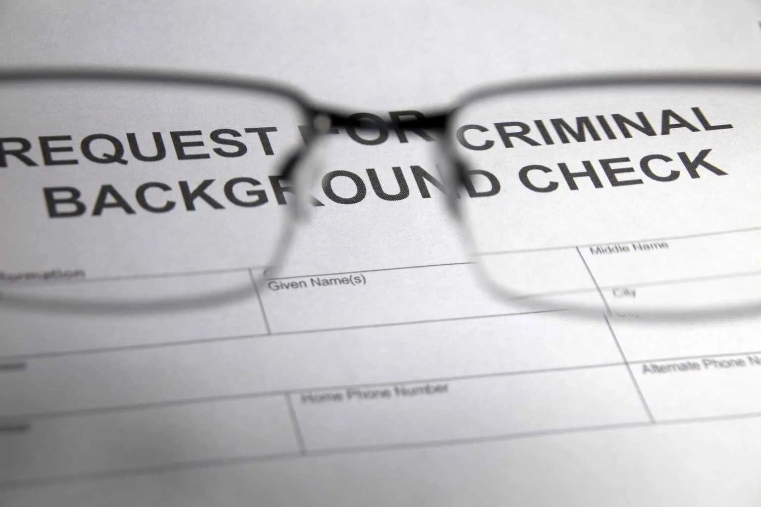 A pair of eyeglasses lays on top of a form with the heading "REQUEST FOR CRIMINAL BACKGROUND CHECK," with the text partially visible through the lenses.