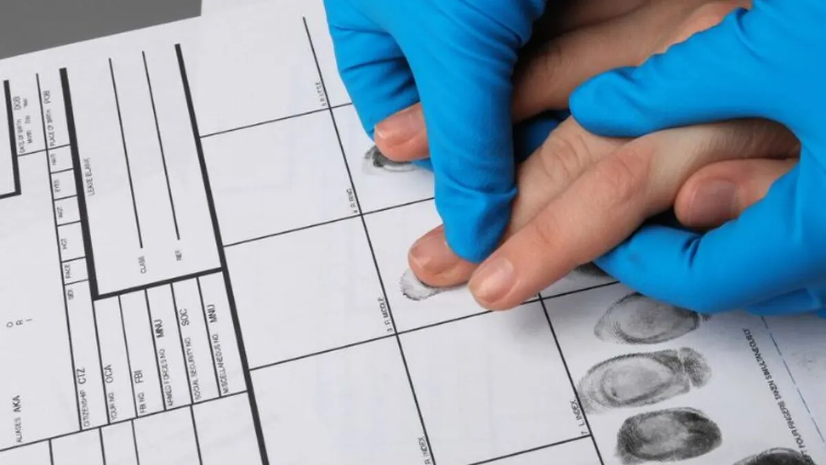 A person wearing blue gloves is carefully pressing another person's fingers onto a fingerprint card to record their fingerprints during a fingerprinting process.