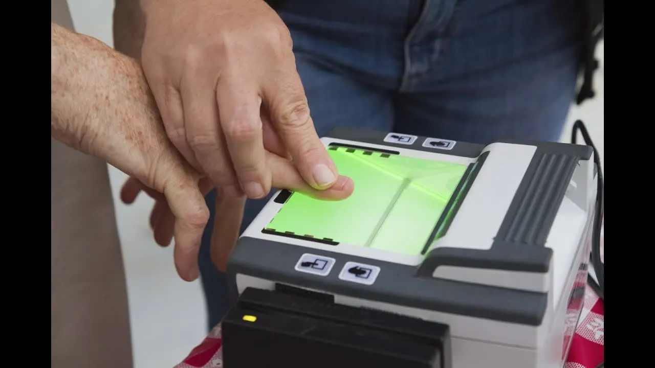 A close-up view of hands pressing a person's fingers onto the glass surface of a biometric fingerprint scanner, capturing fingerprints for identification.